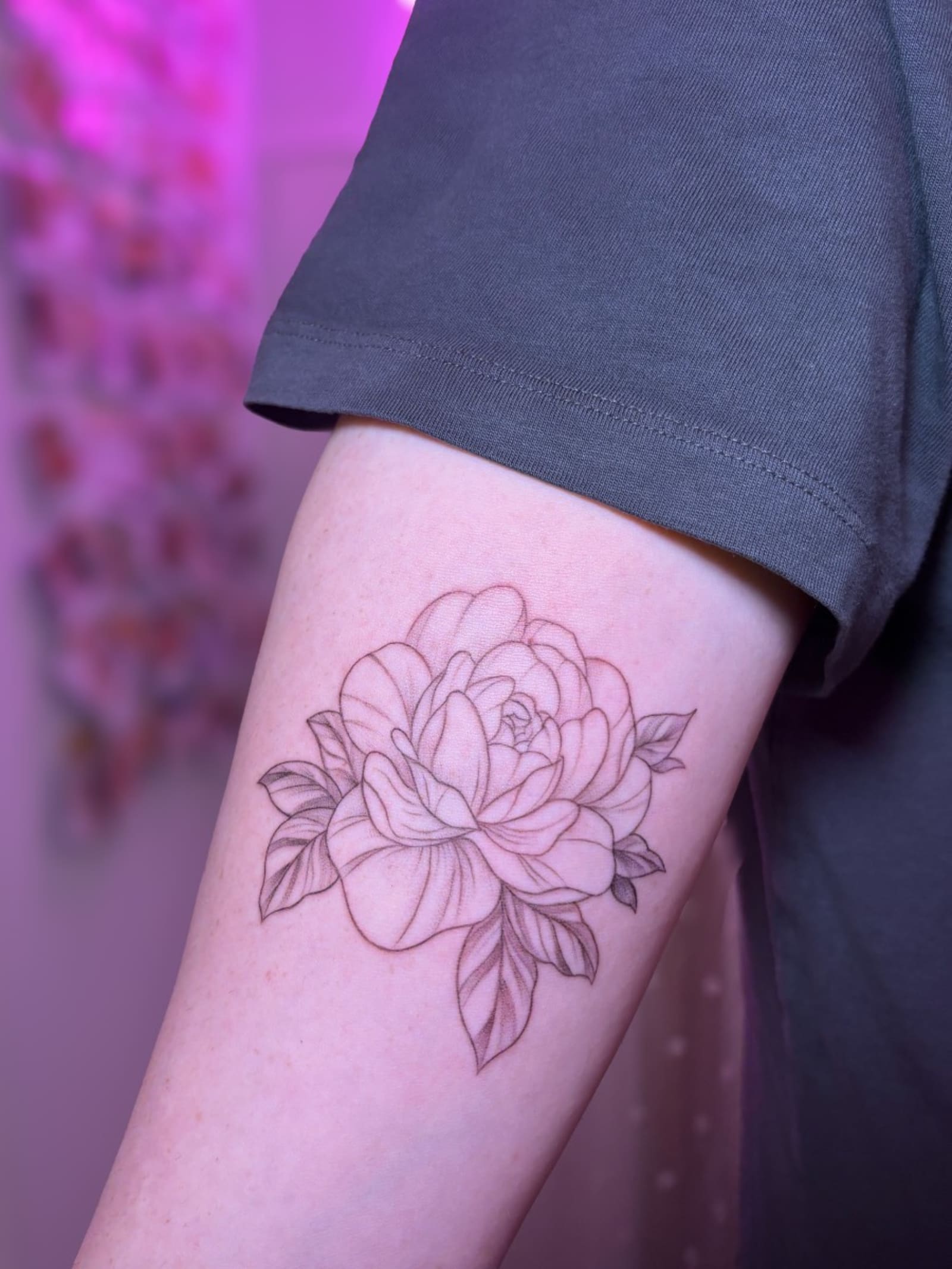 Fine line peony tattoo with shaded leaves on the back of an upper arm